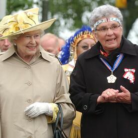 Wearing a beige rain jacket, The Queen walks beside an Indigenous woman wearing a beaded headband and two medals. 