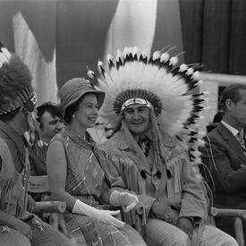 The Queen sits between two Indigenous men wearing ceremonial headdresses.  