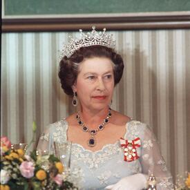 The Queen, wearing a formal dress, the Order of Canada insignia and a jeweled tiara, sits at a table. The table is draped with cloth, and a flower arrangement is in the foreground. 
