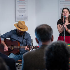 A woman sings into a microphone while a man plays guitar.