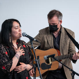 Une femme chante dans un micro tandis qu’un homme joue de la guitare.