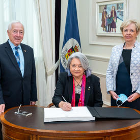 Governor General Mary Simon is seated at a desk. She has a pen in hand and she is signing a guest book. A man is standing to her left and a woman is standing to her right. They are all smiling.