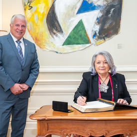 Governor General Mary Simon is seated at a desk. She has a pen in hand and she is signing a guest book. A man is standing to her left. They are both smiling. 