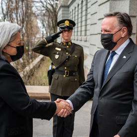 Governor General Mary Simon is shaking hands with the Premier of Quebec. They are outdoors and they are both wearing masks. A guard is standing at attention in the near distance. 