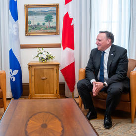 Son Excellence s'entretient avec le premier ministre du Québec. Ils sont tous deux assis et se sourient l'un à l'autre. Le drapeau du Québec et le drapeau canadien sont en arrière-plan.