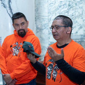 Two men are wearing orange shirts. One is holding a baseball cap.