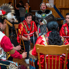 A group of 5 people singing and beating a drum. There are people behind them. They are indoors.