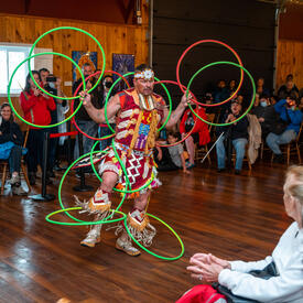A man is performing a dance while handling several hula hoops. There is a crowd around him. They are indoors.