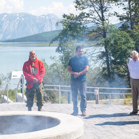 Governor General Simon, Mr. Fraser and two men are standing around a large stone pit. Smoke is rising from the pit. Behind them, there is a lake and a mountain.