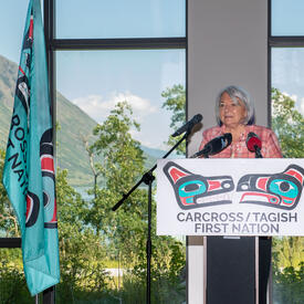 Governor General Simon is standing at a podium. She is speaking into several microphones. A sign on the podium reads, “Carcross/Tagish First Nation.” A large window behind her offers a view of a mountain, trees and a lake.