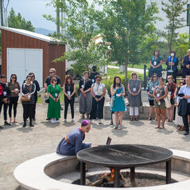 People are standing in a wide circle around a large fire pit.