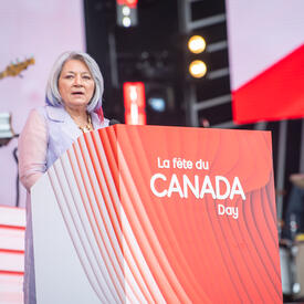 La gouverneure générale est debout derrière un podium ou est inscrit en blanc sur fond rouge “La fête du Canada Day.”