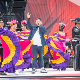 Un artiste chante entouré de danseuses portant des costumes de couleurs pourpre, rose et jaune éclatants sur scène lors de la cérémonie de la fête du Canada.