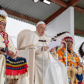Pope Francis is delivering remarks. He is flanked by two Indigenous leaders.
