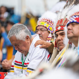 People wearing traditional Indigenous headdresses. A man is bent over with his eyes closed. A man has his hand on his shoulder.