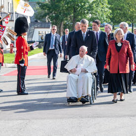 Pope Francis is being wheeled in a wheelchair across the grounds of the GGCitadelle. Governor General Simon, Prime Minister Justin Trudeau, and several others are walking behind him.