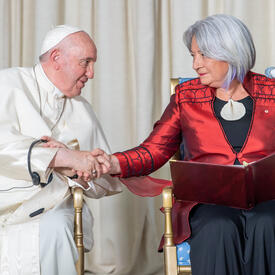 La gouverneure générale Simon est assise à côté de Sa Sainteté le pape François dans une salle du GGCitadelle. Il tient une de ses mains. Elle a un livre ouvert sur ses genoux.