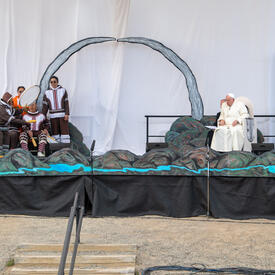 Une grande scène décorée de paysages montagneux. Des artistes chantent et jouent des instruments sur le côté droit de la scène. Le pape François et un autre homme sont assis sur le côté gauche de la scène.