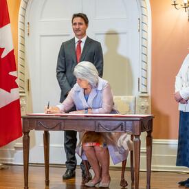 The Governor General is sitting at a desk signing a document. The Prime Minister is standing behind her.