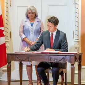 The Prime Minister is sitting at a desk signing a document. The Governor General is standing behind him.