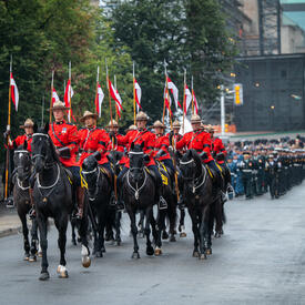 Members of the Royal Canadian Mounted Police ride down a street. The rest of the military parade follows them.