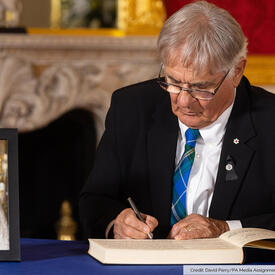 Mr. Whit Fraser is sitting at a table and writing in a book. There is a portrait of Her Majesty The Queen on the table. Text in the lower right corner of the image reads, “Credit: David Parry/PA Media Assignments.”