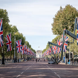 A wide street lined with flags of the United Kingdom. There are people along the left side of the street. Some people are crossing the street.
