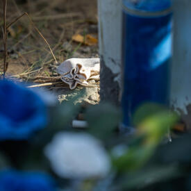 A small tobacco pouch is at the base of a cross. A blue candle and flowers are blurred in the foreground. 