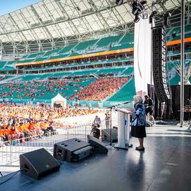 La gouverneure générale Simon est sur une scène dans un stade. Elle se tient derrière un pupitre et parle dans un microphone. Une foule de personnes portant des chandails orange est assise devant la scène.