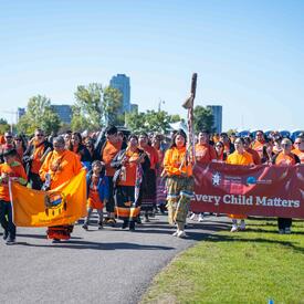 A group of people marching. Many are wearing orange shirts and a large banner that says ‘Every Child Matters’ is visible.