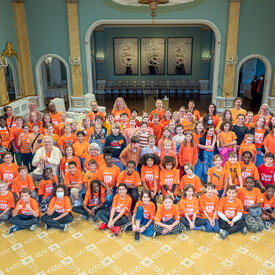 La gouverneure générale Simon et M. Fraser posent pour une grande photo de groupe avec des écoliers dans la salle de bal de Rideau Hall. La plupart des enfants portent un chandail orange.