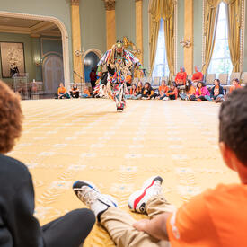 Un danseur se produit dans la salle de bal de Rideau Hall. Il porte une tenue traditionnelle. Des écoliers sont assis autour de lui et le regardent.