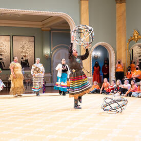 Une danseuse de cerceaux se produisant dans la salle de bal de Rideau Hall. Elle porte une jupe traditionnelle à rubans et tient cinq cerceaux.