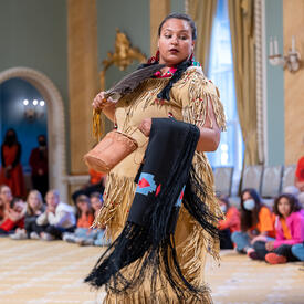 A woman is standing in the Ballroom at Rideau Hall. She is wearing a traditional Indigenous outfit and is holding a feather fan in front of her chest.