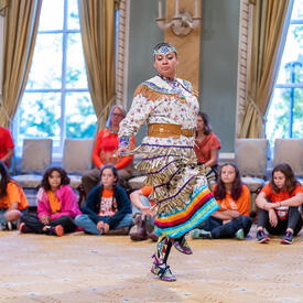 A dancer performing in the Ballroom at Rideau Hall. She is wearing a traditional outfit. Schoolchildren are seated around her, watching.