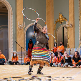 A hoop dancer performing in the Ballroom at Rideau Hall. She is wearing a traditional ribbon skirt and is holding 5 hoops.