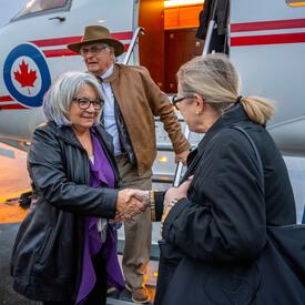 La gouverneure générale serre la main d'une représentante de l'Islande sur le tarmac. M. Fraser descend les marches de l'avion derrière eux.