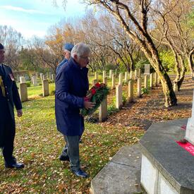 Mr. Fraser, Col. Ronald Walker and a person wearing a military uniform are standing in front of a stone memorial in Fossvogur Cemetery. There is a wreath leaning against the memorial. 