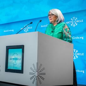 La gouverneure générale se tient derrière un pupitre sur une grande scène. Elle parle dans un microphone. 