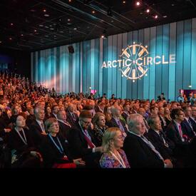 A large seated audience. The wall next to the audience is lit with blue lights and a projection on the wall reads, “Arctic Circle.”