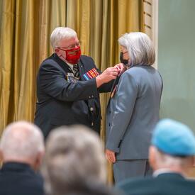 M. Bruce Julian, président national de la Légion royale canadienne, épingle un coquelicot sur la chemise du gouverneure générale Simon. Un public est assis en face d'eux.