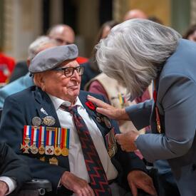 Governor General Simon stands in front of a man who is seated in a wheelchair. She touches a poppy pinned to his shirt. There is a group of people seated around them.