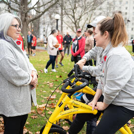 Governor General Mary Simon is speaking with a person who is sitting on a small yellow bicycle. They are in a park. 