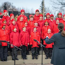 A group of children wearing red coats and hats is singing. They are outside. Someone who has their back to the camera is leading the choir.