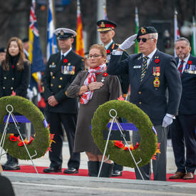 Mme Candy Greff est debout devant deux couronnes de fleurs au Monument commémoratif de guerre du Canada à Ottawa. Un homme est debout à côté d'elle et salue. Plusieurs personnes sont debout derrière eux.