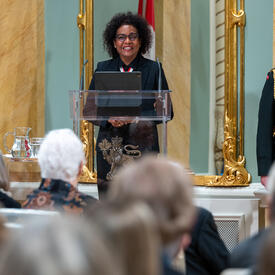 The Right Honourable Michaëlle Jean is standing at a podium is addressing the audience in the Rideau Hall ballroom.