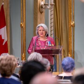 Governor General Mary Simon is wearing a pink suit. She is standing behind a podium in front of a large crowd in the Ballroom of Rideau Hall. A large mirror and a flag of Canada are visible behind her.