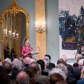 Governor General Mary Simon is standing in front of a large crowd in the Ballroom of Rideau Hall. A large mirror and a large abstract painting are hanging on the wall behind her.