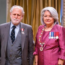La gouverneure générale Simon se tient à côté d’un homme qui porte une médaille épinglée à son blazer gris.
