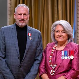Governor General Simon is standing next to a man who has a medal pinned to his shirt blue blazer.